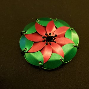 May include: A green and red metal flower-shaped object with a black center. The object is made of many small, overlapping pieces.