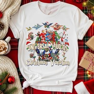 May include: White t-shirt featuring a colorful graphic with the text "UNIVERSAL STUDIOS Christmas Vacation." The design includes various cartoon characters in holiday attire, creating a festive and fun theme.