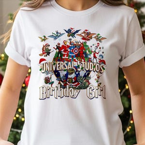 May include: White t-shirt featuring a colorful graphic with the text "UNIVERSAL STUDIOS Birthday Girl." The graphic includes various cartoon characters in Santa hats and Christmas decorations. The shirt has short sleeves.