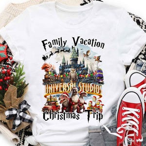 May include: White t-shirt with a graphic of a family vacation to Universal Studios during Christmas. The graphic features a castle, dinosaurs, and Santa Claus. The text "Family Vacation Universal Studios Christmas Trip" is printed on the shirt.