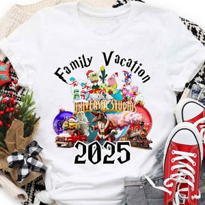 May include: White t-shirt with a graphic of cartoon characters from Universal Studios, a Christmas tree, and the text "Family Vacation 2025".