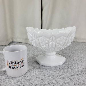 May include: A white milk glass bowl with a scalloped edge and a pedestal base. The bowl has a diamond pattern and is sitting on a gray surface next to a white mug with the text "Vintage Bounce" on it.