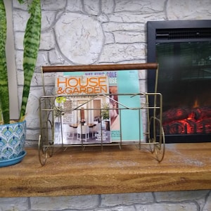 May include: A gold metal magazine rack with a wooden handle holds a copy of House & Garden magazine. The magazine rack is sitting on a wooden mantel above a faux stone fireplace.