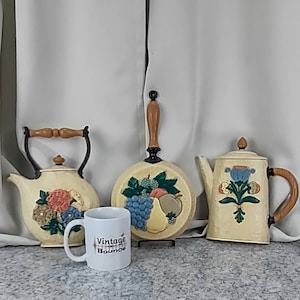 May include: Three vintage kitchenware items with a floral and fruit design. A white teapot with a brown handle, a white coffee pot with a brown handle, and a white frying pan with a brown handle. The frying pan has a design of grapes, a pear, and an apple. The teapot and coffee pot have floral designs. The items are on a white background.