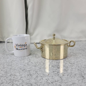 May include: A gold-colored metal saucepan with a lid and two handles. The saucepan is sitting on a white countertop.