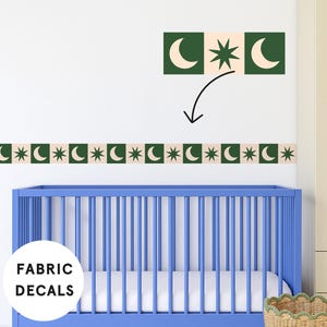 Op de afbeelding: Stofstickers met een herhalend patroon van groene vierkanten, elk met een wit maansikkel- en sterontwerp. De stickers worden boven een blauwe wieg getoond. De woorden "FABRIC DECALS" staan in een witte cirkel.