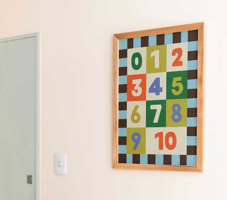 May include: A framed print featuring numbers 0-10 in a colorful grid pattern. The numbers are in various colors, including green, orange, and blue, set against a checkered background. The frame is made of light-colored wood.