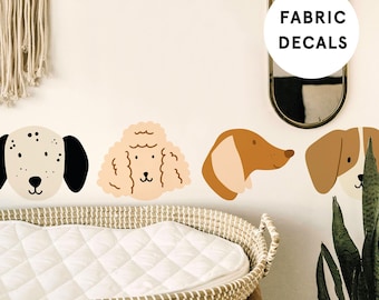 Op de afbeelding: Een set van vier stoffen wanddecoraties met verschillende hondenrassen. De decoraties zijn in een rij op een witte muur. De decoraties zijn in cartoon-stijl en tonen een dalmatiër, een poedel, een teckel en een beagle. De tekst "FABRIC DECALS" staat in een cirkel boven de decoraties.