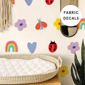 May include: A set of colourful fabric decals featuring a rainbow, hearts, flowers, ladybirds, and a butterfly. The decals are shown on a white wall with a wicker bassinet in the foreground. The text "FABRIC DECALS" is visible in the top right corner of the image.