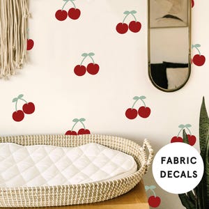 May include: A white changing mat with a wicker basket underneath. The wall behind the mat is decorated with red cherry fabric decals. The text "FABRIC DECALS" is visible in the lower right corner of the image.