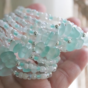 May include: A stack of delicate bracelets made with small, light blue and white glass beads. The bracelets are layered on top of each other, creating a beautiful and intricate design.