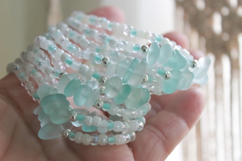 May include: A stack of delicate bracelets made with small, light blue and white glass beads. The bracelets are layered on top of each other, creating a beautiful and intricate design.
