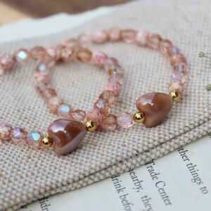 May include: Two pink and brown beaded bracelets with heart-shaped charms. The bracelets are on a brown fabric surface.