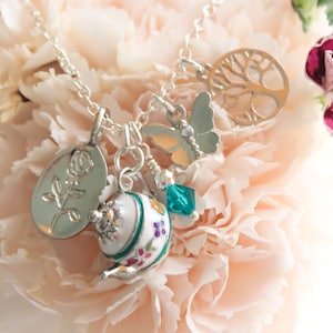 May include: A silver necklace with multiple charms, including a teapot, a rose, a butterfly, and a tree of life. The charms are hanging from a silver chain and are displayed against a background of pink carnations.