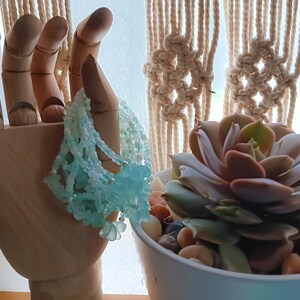 May include: A wooden hand model holding a light blue beaded necklace with a flower design. The hand is in front of a macrame wall hanging and a potted succulent plant.