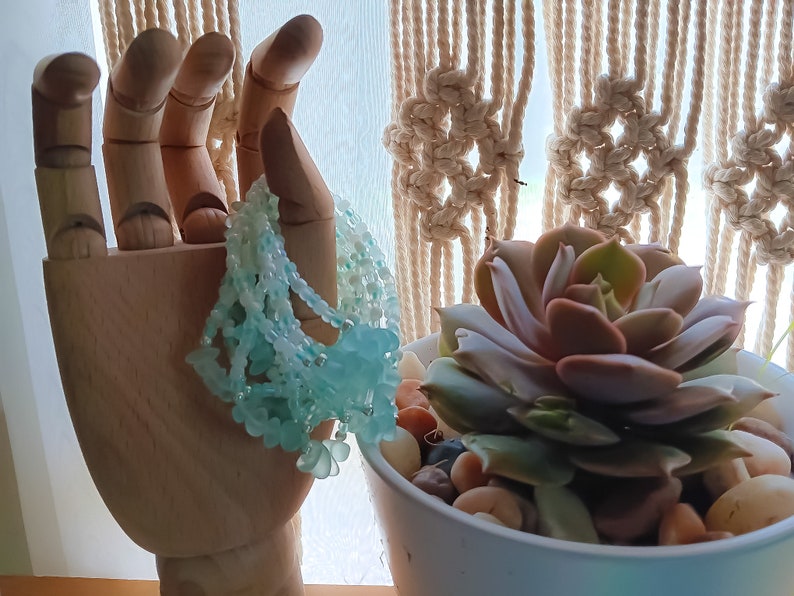 May include: A wooden hand model holding a light blue beaded necklace with a flower design. The hand is in front of a macrame wall hanging and a potted succulent plant.