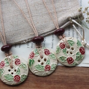 May include: Three ceramic button necklaces with a floral design. Each necklace features a white button with a red rose and green leaf design. The necklaces are strung on brown leather cord with dark red beads.