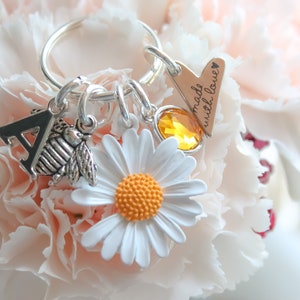 May include: A silver keychain with a charm that says "made with love" and a heart, a silver letter A charm, a silver bee charm, a white daisy charm with an orange center, and a yellow crystal charm.