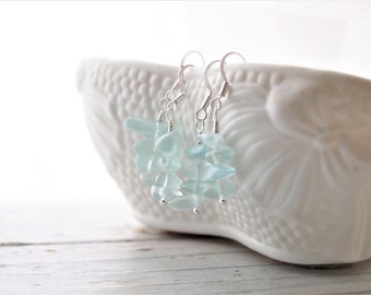 May include: A pair of silver earrings with light blue glass beads. The earrings are hanging from a white bowl.