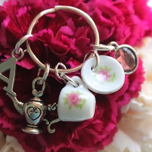 May include: A silver charm keychain with a teacup, teapot, and a letter A charm. The charms are attached to a silver ring. The keychain is surrounded by pink and white carnations.