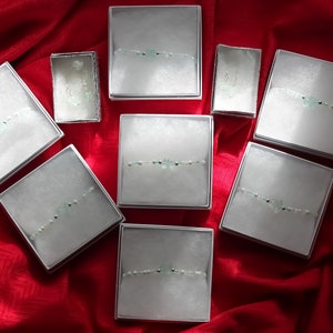 May include: Ten small white boxes with lids, each containing a delicate bracelet with light green stones and silver beads. The boxes are arranged in a heart shape on a red fabric background.