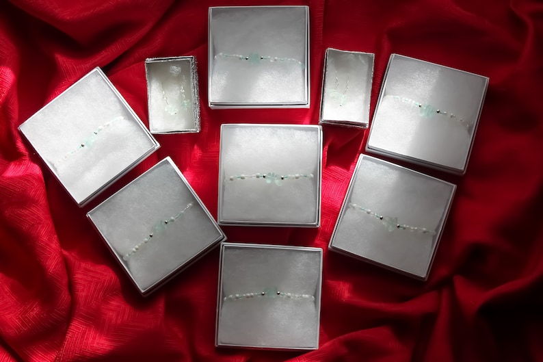 May include: Ten small white boxes with lids, each containing a delicate bracelet with light green stones and silver beads. The boxes are arranged in a heart shape on a red fabric background.