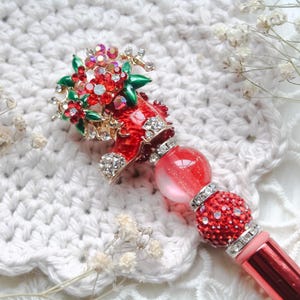 May include: A red and gold pen with a decorative top featuring a red shoe adorned with rhinestones and a floral arrangement. The pen has a red barrel and a red and pink jewelled accent.
