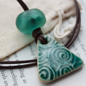 May include: A teal blue ceramic pendant with a swirling pattern hangs from a brown leather cord. The pendant is attached to a teal blue glass bead.