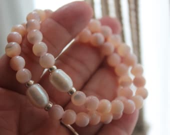 Puede incluir: Dos pulseras hechas de cuentas rosadas y blancas. Las pulseras están superpuestas y tienen un acabado nacarado.