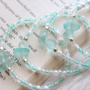 May include: A set of three bracelets with white and light blue beads. The bracelets feature light blue sea glass charms.