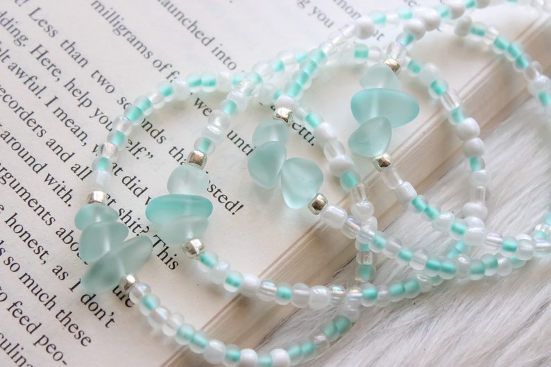 May include: A set of three bracelets with white and light blue beads. The bracelets feature light blue sea glass charms.