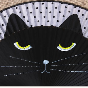 May include: A black folding fan with a white and black polka dot design on the top half. The bottom half of the fan is painted with a black cat face with yellow eyes and a white nose.