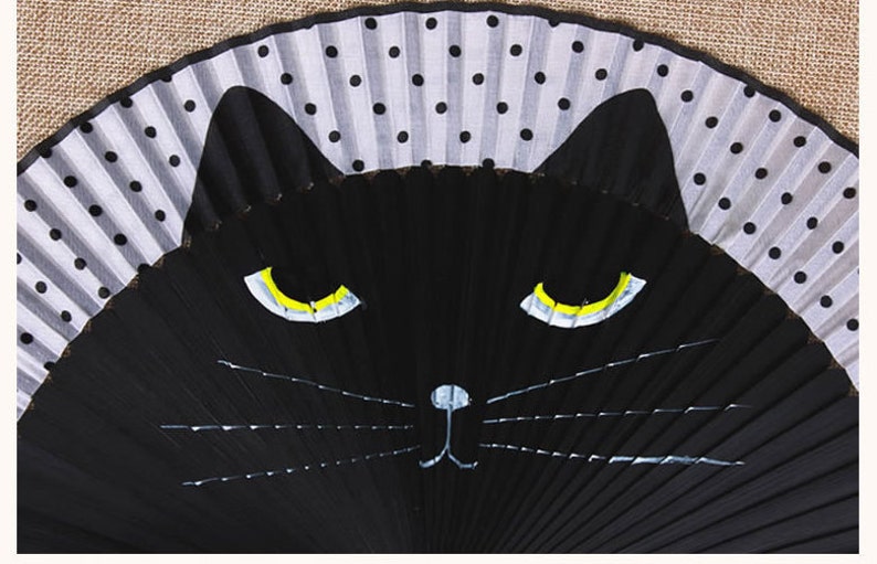 May include: A black folding fan with a white and black polka dot design on the top half. The bottom half of the fan is painted with a black cat face with yellow eyes and a white nose.