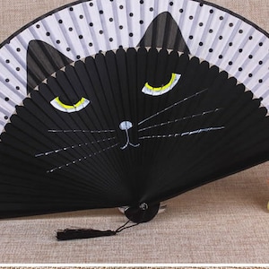 May include: A black folding fan with a white and black polka dot pattern. The fan has a black cat face painted on it with yellow eyes and a white nose.