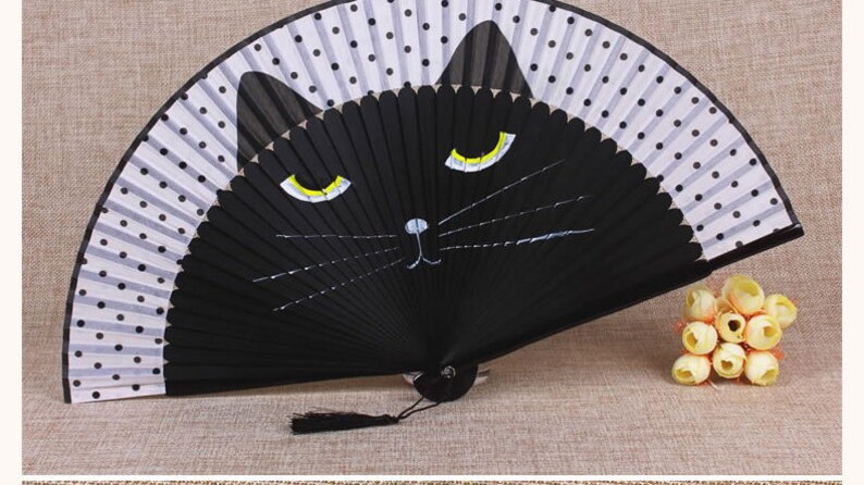 May include: A black folding fan with a white and black polka dot pattern. The fan has a black cat face painted on it with yellow eyes and a white nose.