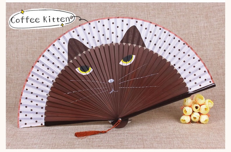 May include: A brown folding fan with a white background and black polka dots. The fan has a painted image of a brown cat with yellow eyes. The fan has a red tassel.