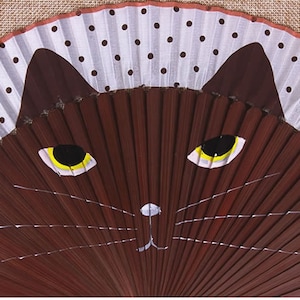 May include: A brown folding fan with a white and black polka dot design. The fan is painted with a brown cat face with yellow eyes and white whiskers.