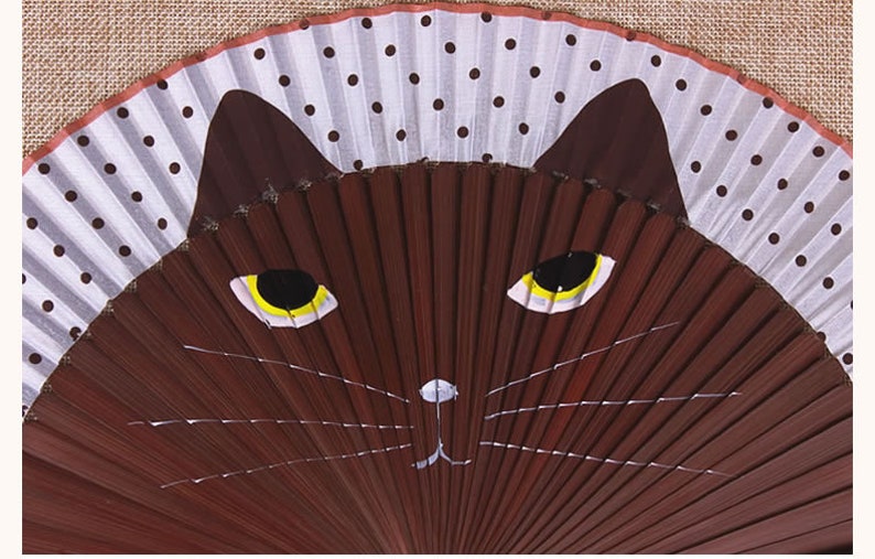 May include: A brown folding fan with a white and black polka dot design. The fan is painted with a brown cat face with yellow eyes and white whiskers.