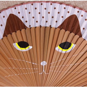 May include: A brown and white folding fan with a painted cat face. The cat has yellow eyes and a white nose.