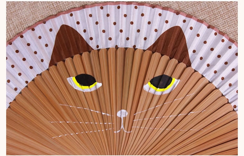 May include: A brown and white folding fan with a painted cat face. The cat has yellow eyes and a white nose.