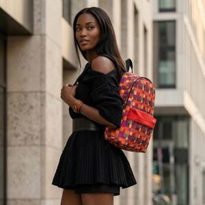 Puede incluir: Una mochila colorida con un patrón geométrico de cuadrados naranjas, rojos, morados y amarillos. La mochila tiene un bolsillo frontal rojo y correas negras. La persona lleva un atuendo negro.