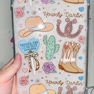 Puede incluir: Funda de teléfono transparente con temática western. El diseño incluye sombreros de vaquero, botas, herraduras, cactus, rosas y los textos "Howdy Darlin'" y "Sucker for Cowboy Romance". La funda está decorada con corazones y estrellas.