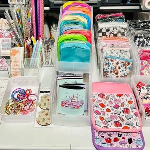 Puede incluir: Exhibición de varios accesorios y artículos de papelería. Incluye pegatinas, gomas para el pelo, bolsas y enfriadores de bebidas. Los artículos están dispuestos en recipientes de plástico transparente y sobre una superficie blanca.