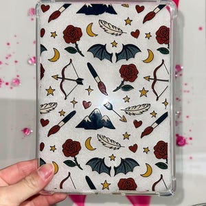 Puede incluir: Funda protectora transparente con fondo blanco con un patrón repetido de rosas rojas, murciélagos, arcos y flechas, pinceles, estrellas, lunas y plumas. La funda está sujeta con una mano.