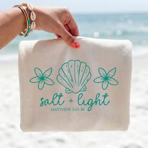 Op de afbeelding: Een crèmekleurig canvas tasje met een groenblauwe afbeelding van een schelp geflankeerd door twee bloemen. Onder de afbeelding staat de tekst "salt + light" in een cursief lettertype, gevolgd door "MATTHEW 5:13-16" in een eenvoudig lettertype.