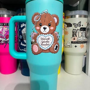 May include: A matte turquoise tumbler with a handle and a lid, featuring a brown teddy bear illustration holding a heart-shaped sign that reads "Dearest Gentle Reader" surrounded by small flowers, a bee, and a light blue bow on its head.