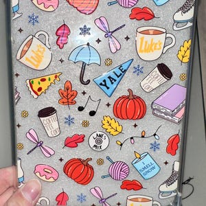 Puede incluir: Funda de teléfono transparente con un fondo gris y gráficos coloridos con temática otoñal. El diseño incluye calabazas, hojas, tazas de café, rosquillas y el texto "Luke's" y "I Smell Snow".