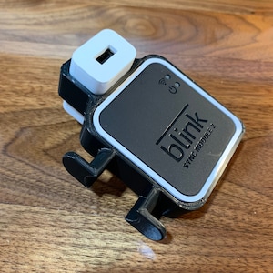 Può includere: Un modulo di sincronizzazione Blink 2 nero e bianco con una porta USB bianca nella parte superiore. Il modulo ha un bordo bianco e la parola "blink" in lettere bianche.