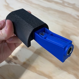 May include: A blue and black woodworking jig with a small metal drill bit. The jig is designed to help with pocket hole joinery.