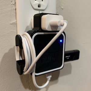 Puede incluir: Un soporte de pared negro con un adaptador de corriente blanco enchufado a una toma de corriente. Un cable de carga blanco está envuelto alrededor del soporte y un dispositivo negro con el texto "blink" está conectado al soporte. Una unidad USB negra está conectada al dispositivo.